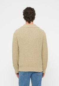 Mężczyzna z kręconymi brązowymi włosami, ubrany w beżowy, dziergany sweter i niebieskie dżinsy, widziany od tyłu na jednolitym białym tle.