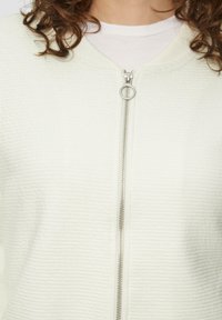 Šviesiai kreminė zip-up striukė iš tekstūruotos medžiagos, turinti sidabrinį užtrauktuką ir apvalų traukimo elementą. Apykaklė yra minkšta, bet struktūruota.