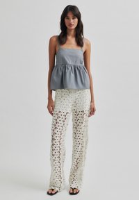 Top peplum grigio con spalline regolabili, abbinato a pantaloni bianchi in pizzo lavorato a uncinetto con motivi floreali e silhouette a gamba larga. Sandali neri.