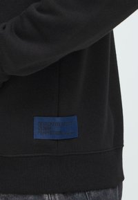 Primo piano di una felpa nera con un'etichetta blu vicino all'orlo che recita "Dedicate alla Perfezione del Denim" e parte di un braccio di persona e jeans visibili.
