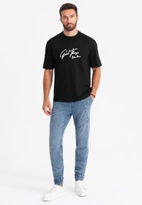 Camiseta negra de manga corta con texto en cursiva blanca que dice "Buenas Cosas" en la parte frontal, combinada con pantalones de chándal azul claro y zapatillas blancas.