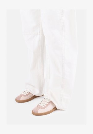 Hellrosa und beige Sneaker mit weißen Schnürsenkeln, kombiniert mit weit geschnittenen weißen Hosen mit sichtbarer Naht und geradem Schnitt.