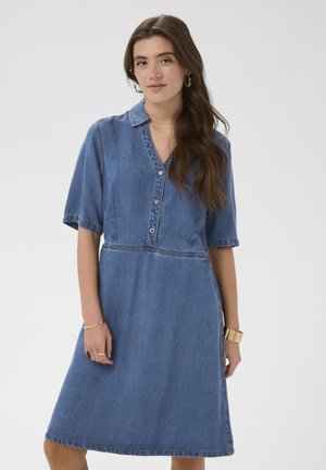 Jeune femme aux longs cheveux bruns portant une robe en jean bleue longueur genou à manches courtes et des boucles d'oreilles créoles dorées.