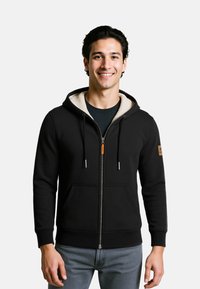 Felpa nera con zip, cappuccio foderato in pile crema, tasche a marsupio, polsini a coste e un dettaglio arancione sulla cerniera.