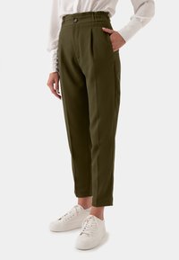 Olijfgroene, hoge broek met plooien, zijzakken en een knoopsluiting. Gestyled met witte sneakers voor een casual look.