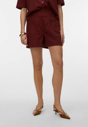 Vrouw draagt hooggetailleerde bordeauxrode korte broek en bijpassend overhemd met knopen, gecombineerd met bruine puntige slingback-hakken en gouden armbanden.