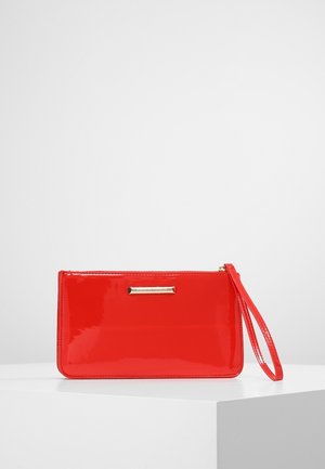 Lesklá červená zápěstní taštička se zlatou destičkou se jménem a zipem, umístěná na bílém podstavci na světle šedém pozadí.