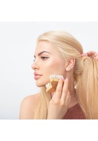 Drewniana szczotka do włosów z miękkim białym włosiem, trzymana w dłoni blisko twarzy osoby, która ma długie blond włosy i gładką cerę.