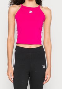 Fuchsia träningscrop-top med tunna band, med vitt Adidas-logotyp och sidostråkar. Matcher med svarta byxor med logotyp på höften.
