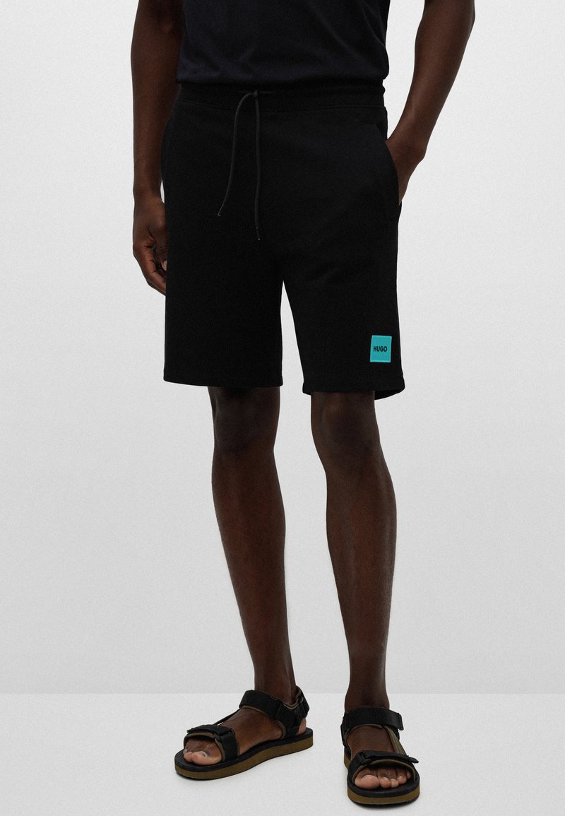 Svarte bomullsshorts med elastisk midjebånd og snor. Har et lite turkist merke på venstre ben. Kombinert med svarte sandaler.