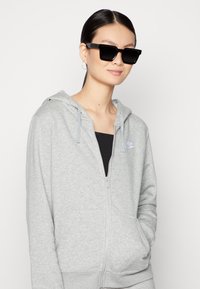 Siva zip-up kapuca iz mehkega materiala z kenguru žepom, v kombinaciji z črnimi kvadratnimi sončnimi očali in črnim vrhom pod njim.