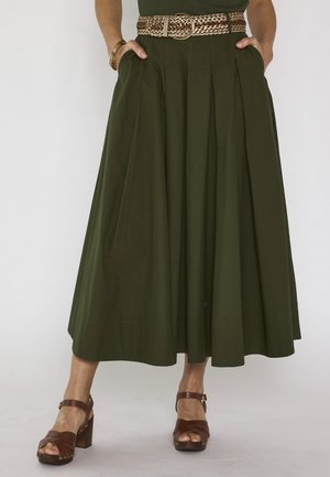 Donna con gonna midi plissettata verde oliva, cintura intrecciata color cuoio e sandali marroni con tacco, mani nelle tasche, in piedi su pavimento grigio.