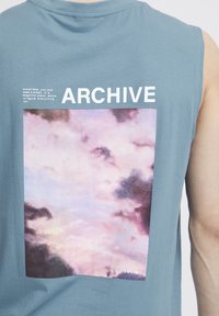 Maglietta senza maniche di colore azzurro chiaro con una stampa grafica di nuvole nelle sfumature di rosa chiaro e grigio, con il testo "ARCHIVE" sopra l'immagine.