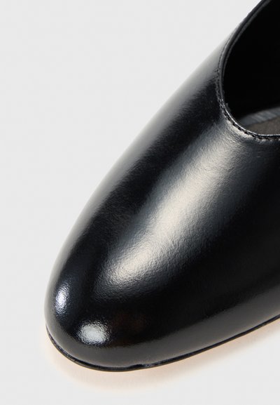 Chaussure en cuir noir avec une finition brillante, bout arrondi et design découpé sur les côtés, mettant en valeur une texture lisse et un style minimaliste.