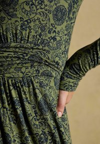 Zbliżenie na talię i ramię osoby ubrane w oliwkowozieloną sukienkę w ciemnoniebieskie wzory kwiatowe, ręka oparta na biodrze.