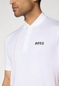 Weißes Poloshirt mit Slim Fit, versehen mit einem schwarzen "BOSS"-Logo, einem knöpfbaren Kragen und diagonalen Streifen in hellen Farben auf einer Seite.