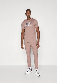 Világosbarna sportos póló "New Balance" logóval, világosbarna melegítőnadrággal és fekete-fehér sportcipőkkel párosítva. Simább textúra, testhezálló szabás.