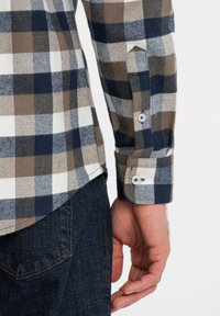 Camicia a quadri con quadrati blu navy, marroni e bianchi. Tessuto di cotone con polsini abbottonati e orlo curvato. Abbinata a jeans scuri.