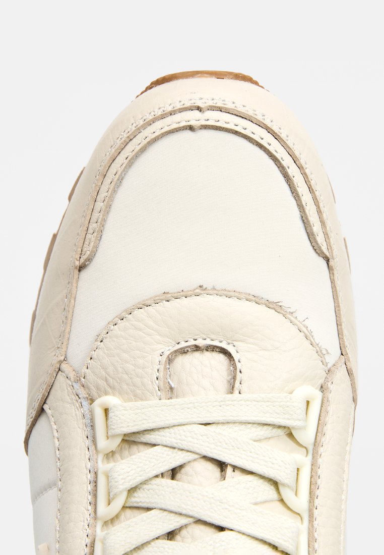 Zapatillas de cuero beige con paneles lisos y texturizados, que presentan una puntera redondeada, cordones planos y una suela de goma. Costuras visibles a lo largo de los bordes.