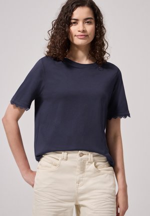Mujer con pelo rizado que lleva una camiseta azul oscuro de manga corta con ribete de encaje y pantalones beige de tiro alto, de pie con una mano en la cadera.