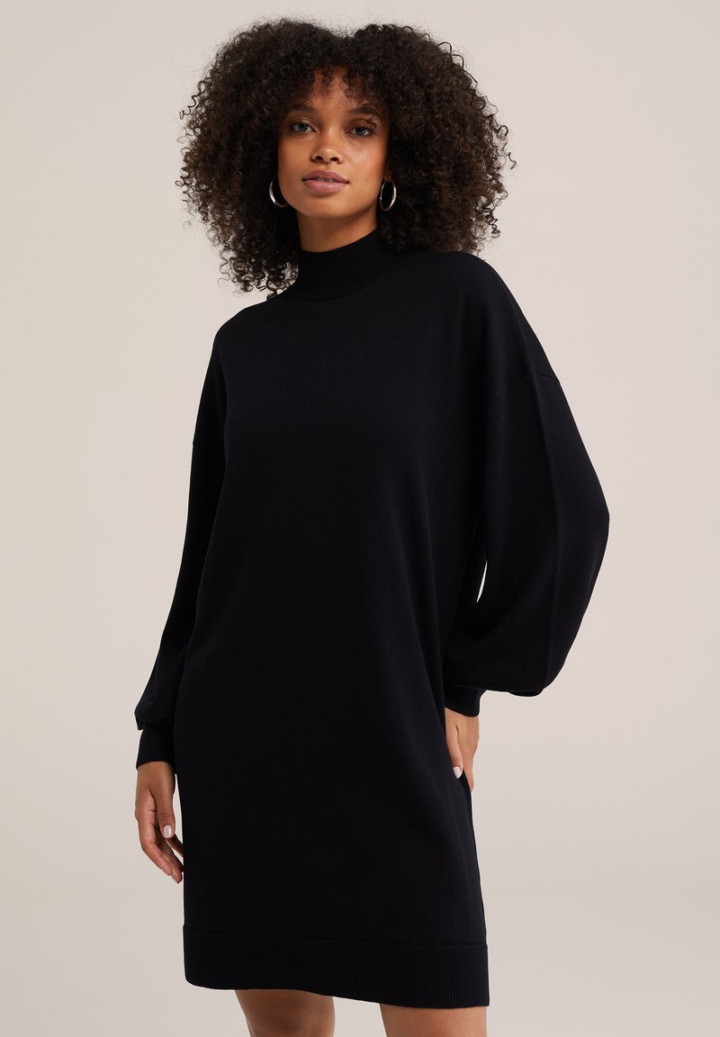 Femme aux cheveux bouclés portant une robe-pull noire à manches longues avec un col montant, debout devant un fond clair uni.