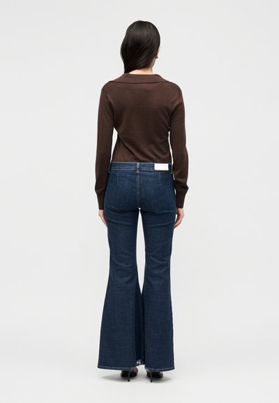 Γυναίκα με μακριά σκούρα μαλλιά, φορώντας καφέ μπλούζα με μακριά μανίκια και σκούρο μπλε καμπάνες τζιν, στέκεται γυρισμένη πλάτη σε απλό φόντο.