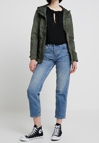 Grøn letvægtsjakke med hætte, sort skjorte med nøglehul-udskæring, lyseblå denimjeans og sorte høje sneakers.