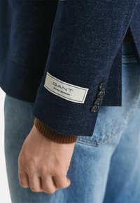 Marineblaue Wolljacke mit einem Markenlabel und vier schwarzen Knöpfen am Ärmel. Gerippter brauner Strick am Handgelenk. Strukturiertes Stofffinish.