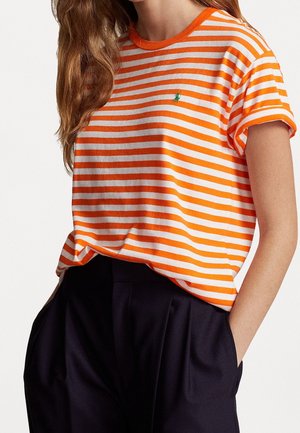 Kobieta w pomarańczowo-białej koszulce w poziome paski z krótkim rękawem z małym zielonym logo, do tego czarne spodnie z wysokim stanem, ręce w kieszeniach.