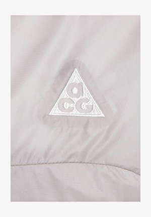 Vaalean taupe -kankaan sileä pinta, jossa on valkoinen brodeerattu kolmion muotoinen logo, jossa on pyöreät korostukset keskellä.