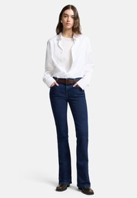 Witte blouse over een beige top, gecombineerd met donkerblauwe flared jeans en een bruine riem. Zwarte enkellaarsjes maken de outfit compleet.