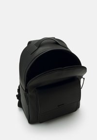 Zaino in pelle nera con parte superiore arrotondata, texture liscia, apertura con zip e tasca frontale con una patta rettangolare. Design minimalista.