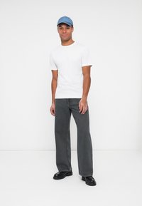 Witte t-shirt met korte mouwen, grijze brede spijkerbroek en een lichtblauw baseballpet. Zwarte chunky schoenen, eenvoudige achtergrond.