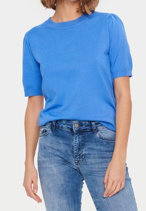 Kvinde iført en kortærmet blå striktrøje med puffede skuldre og lyseblå højtaljede jeans mod en hvid baggrund.