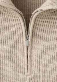 Gros plan d'un pull en maille côtelée beige avec une fermeture éclair argentée partiellement ouverte et un col montant visible.