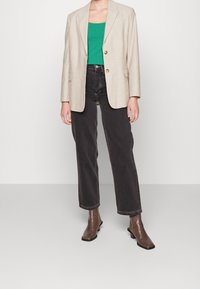 Blazer de lino beige con dos botones y bolsillos delanteros, llevado sobre una camiseta de tirantes verde, combinado con jeans negros de pierna ancha y botas cortas marrones.