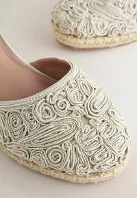 Gros plan sur une espadrille beige ornée d'une broderie complexe en corde blanche avec des motifs tourbillonnants et linéaires.