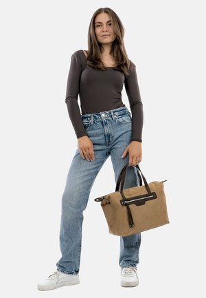 Brown canvas tote bag with black accents, zipper, and dual handles. Model wears fitted long-sleeve top and light blue jeans. White sneakers.