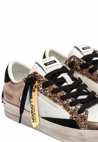Sneakers bianchi e neri con accenti dorati glitterati, dotati di lacci neri, suola nera e etichette di branding sui lati.