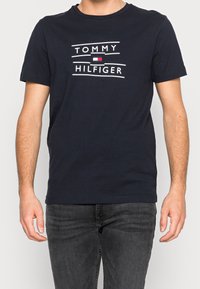 Marineblå bomulds T-shirt med et centralt hvidt logo med "TOMMY" ovenfor og "HILFIGER" nedenfor, sat mod et enkelt linjært design.