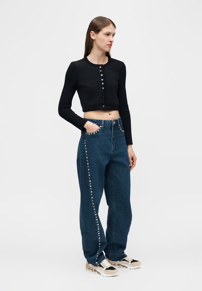 Νεαρή γυναίκα με μαύρο κοντό ζακέτα με κουμπιά, ψηλόμεσο τζιν με τρουκς και μπεζ αθλητικά παπούτσια, στέκεται με το ένα χέρι στην τσέπη.