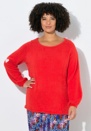 Kvinde med krøllet hår iført en lys rød, fuzzy sweater og et farverigt blomsterprintet skørt, stående med den ene hånd på hoften mod en hvid baggrund.