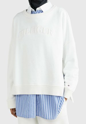 Sweat-shirt blanc oversize avec texte en relief "HILFIGER" porté sur une chemise à rayures verticales bleues et blanches et un foulard noir.