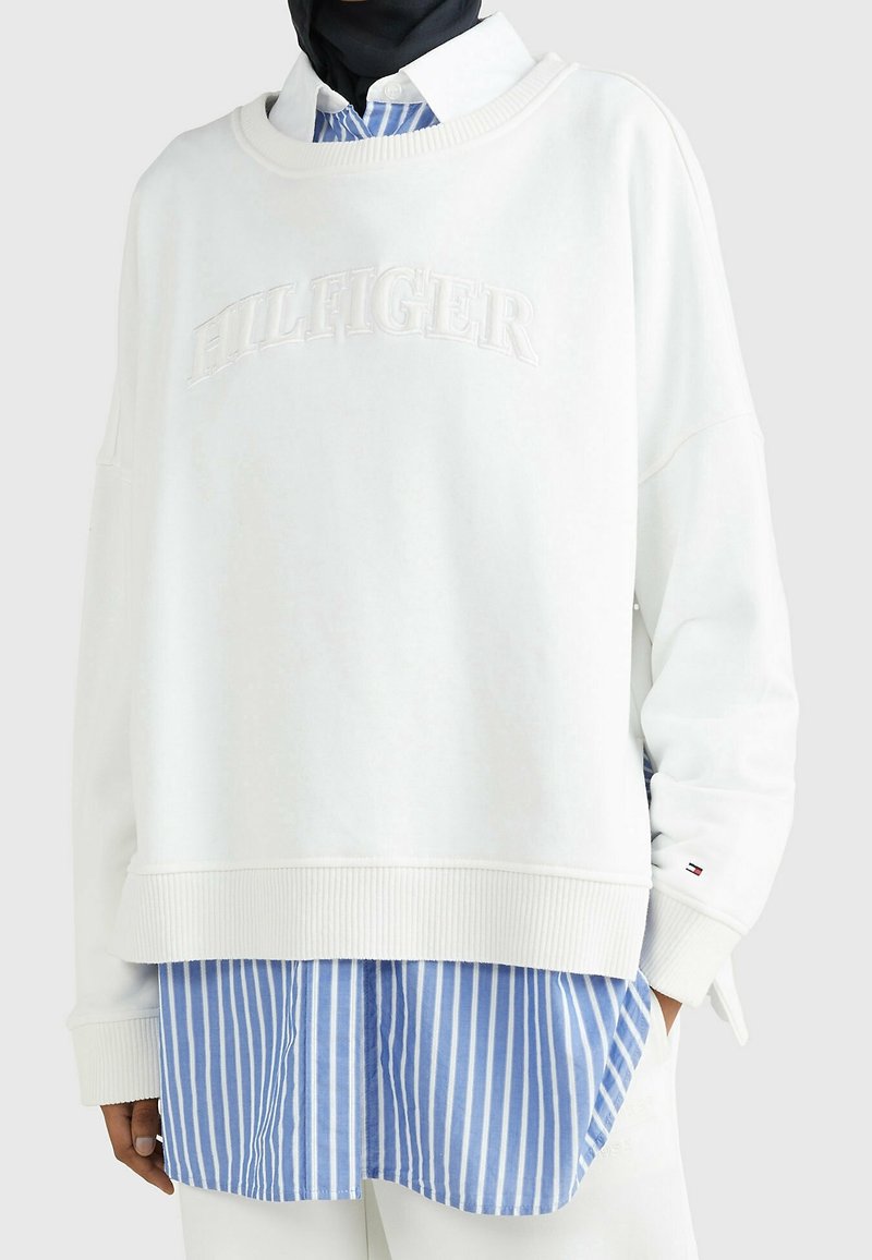 Sweat-shirt blanc oversize avec texte en relief "HILFIGER" porté sur une chemise à rayures verticales bleues et blanches et un foulard noir.