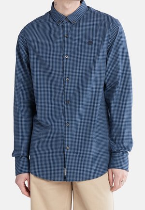 Mann trägt ein blau-schwarz kariertes Hemd mit langen Ärmeln und beigen Hosen, steht vor einem schlichten Hintergrund.