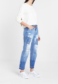 Jersey blanco texturizado, jeans azules desgastados con parches y tachuelas, rematados con zapatillas blancas con detalles decorativos.