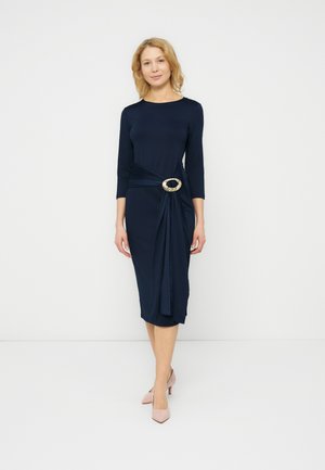 Kvinde iført en navyblå midikjole med trekvart lange ærmer og en dekorativ sølvspænde i taljen, kombineret med lyserøde hæle.