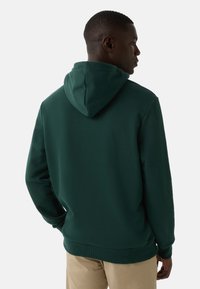 Mężczyzna w ciemnozielonym hoodie i beżowych spodniach, stojący plecami do widza, zwrócony w prawą stronę na jednolitym tle.
