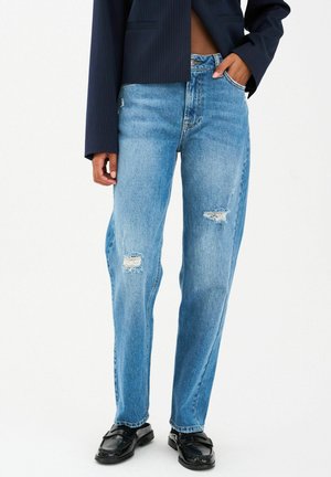 Osoba nosi svetlomodré roztrhané rifle, čierne mokasíny a tmavomodré skrátené sako s pásikmi, s jednou rukou v vrecku.