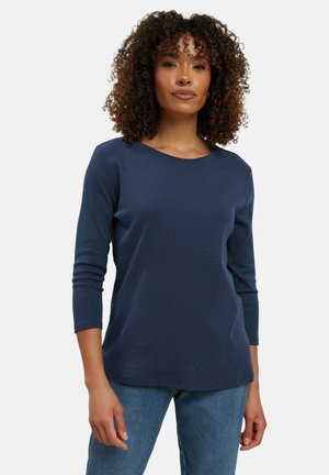 Kvinde med krøllet hår iført en mørkeblå langærmet skjorte og blå jeans, stående foran en ensfarvet lys baggrund.